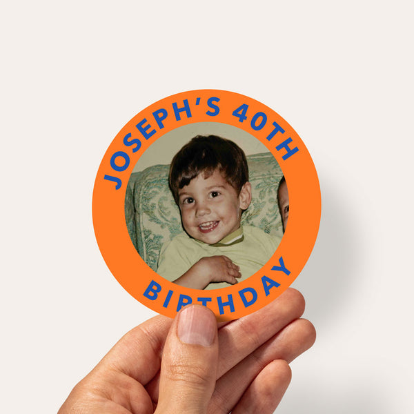 A hand holding a round orange sticker with blue text reading 'JOSEPH'S 40TH BIRTHDAY' and a vintage-style photo of a young boy in the center, against a plain light background.