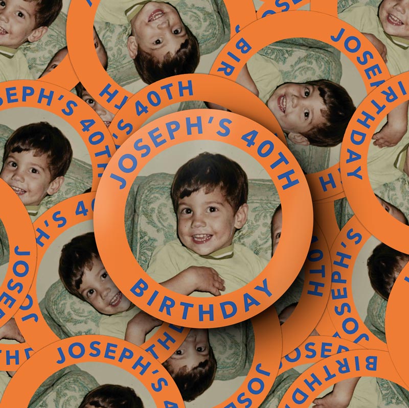 A hand holding a round orange sticker with blue text reading 'JOSEPH'S 40TH BIRTHDAY' and a vintage-style photo of a young boy in the center, against a plain light background.